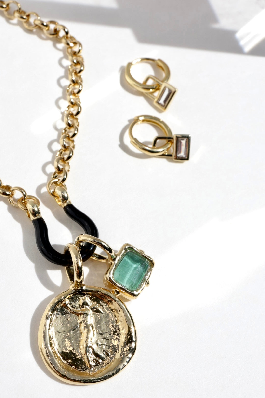 Gold-tone jewelry set featuring a chain necklace with a round coin pendant, a black curved accent, and a green rectangular stone charm, alongside matching small hoop earrings with rectangular clear stones, arranged on a white surface.