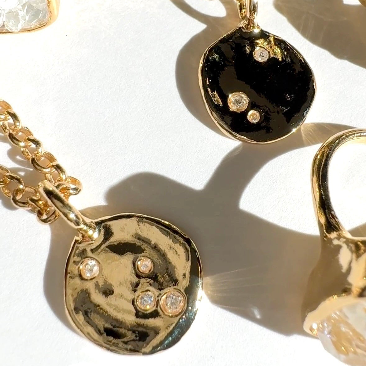 Gold chain necklaces with irregular polished pendants, including one black disc pendant with small clear stones and one gold disc pendant with clear stones, arranged on a white surface with shadows.
