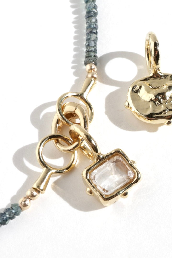 Gold-toned jewelry charm with a rectangular clear gemstone pendant, shown on a white background with part of a matching gold heart-shaped charm and a gray beaded cord visible.