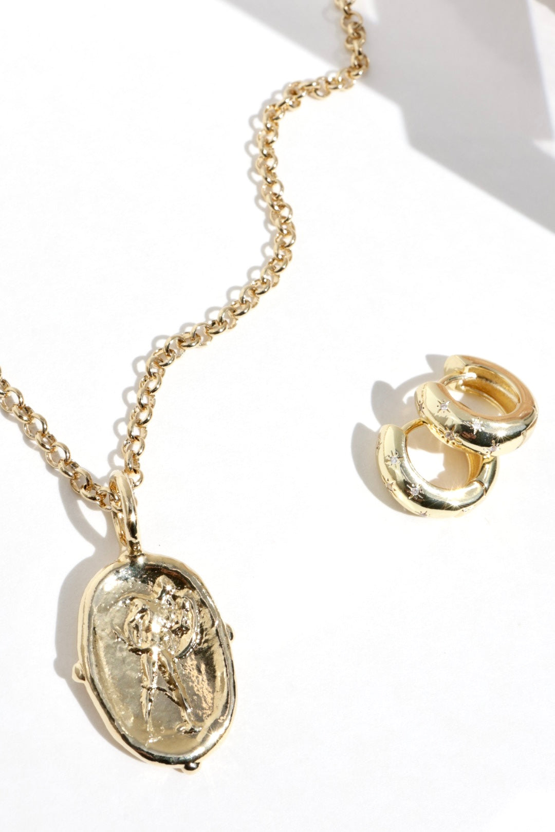 Gold jewelry set featuring an oval pendant necklace with an embossed figure and matching stacked huggie hoop earrings, displayed on a clean white background with soft shadows.