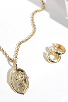 Gold jewelry set featuring an oval pendant necklace with an embossed figure and matching stacked huggie hoop earrings, displayed on a clean white background with soft shadows.