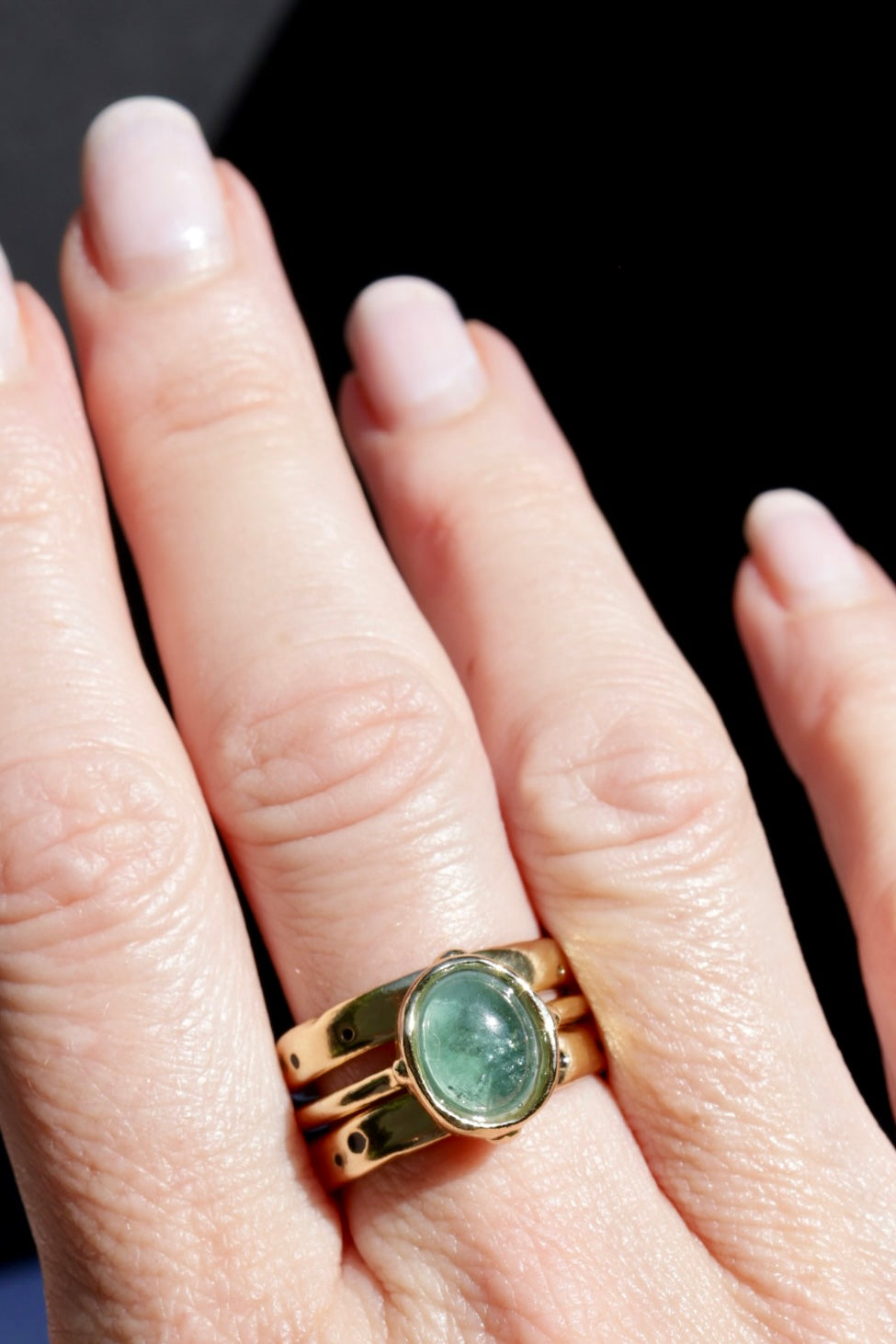 A close-up of a hand wearing a gold ring with an oval green cabochon gemstone, set against a dark background.
